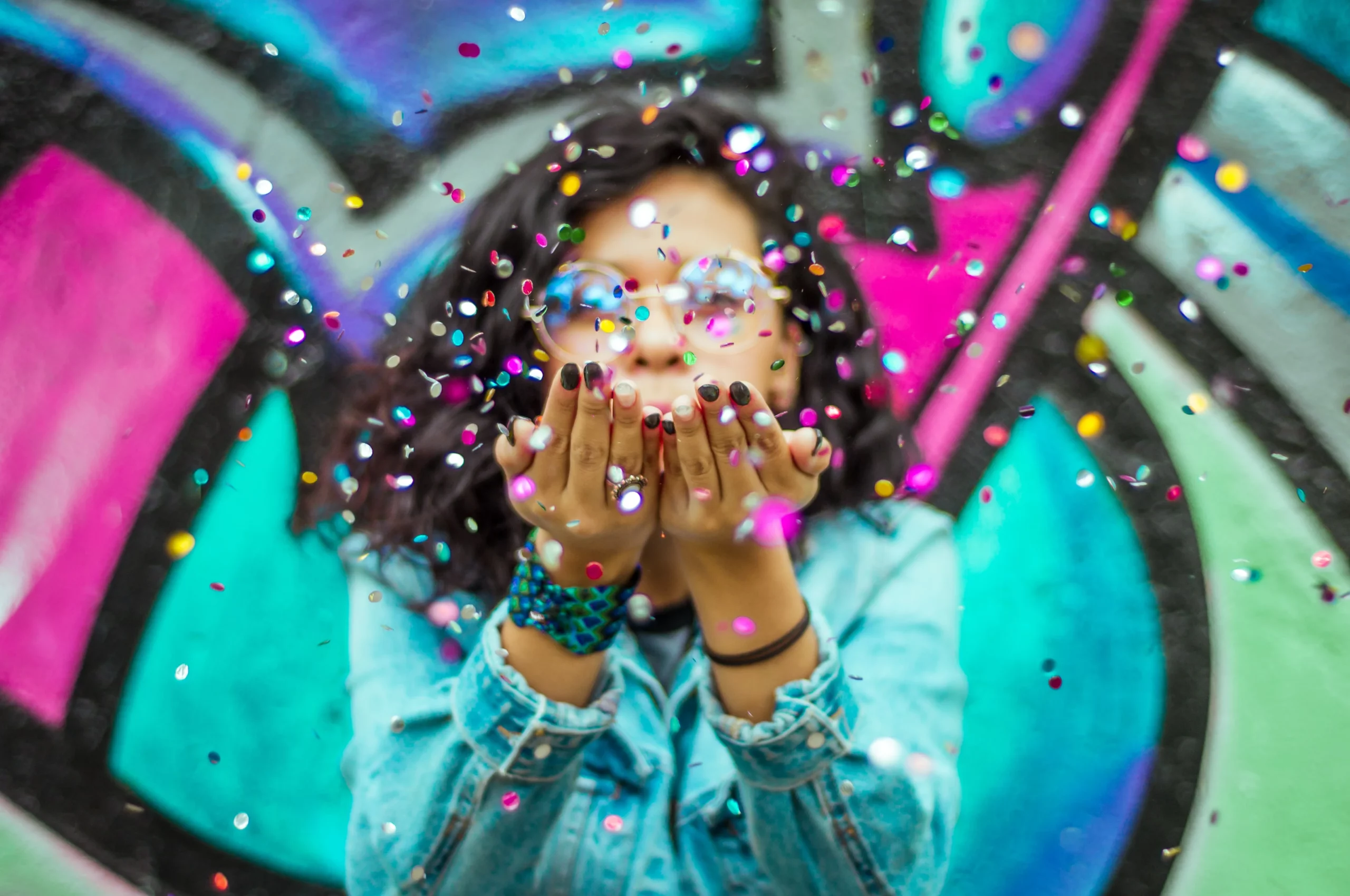 Lady dresses in a blue shirt blowing colourful confettis to the camera.