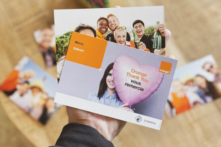 A hand holding two personalised photo postcards. The front postcard shows a woman holding a heart-shaped balloon with the text “Orange Thank You vous remercie” and includes the Stampix and Orange logos. The second postcard behind it features a group of smiling friends holding drinks. Blurred photos are visible in the background on a wooden surface.