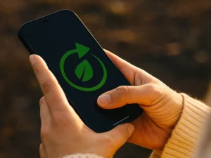 Hands holding a smartphone outdoors, with a green sustainability icon (leaf and circular arrows) displayed on the screen, symbolizing eco-friendly technology and responsible digital connectivity.