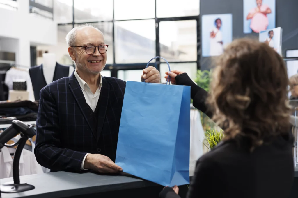 Elderly man smiling while receiving a blue shopping bag from a cashier at a modern retail clothing store counter.