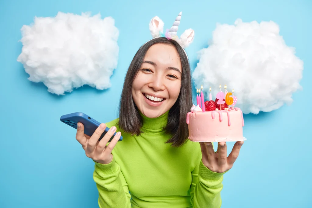 Smiling woman wearing a unicorn headband holds a pink birthday cake with candles showing the number 26 in one hand and a smartphone in the other, standing against a bright blue background with fluffy white cloud decorations.
