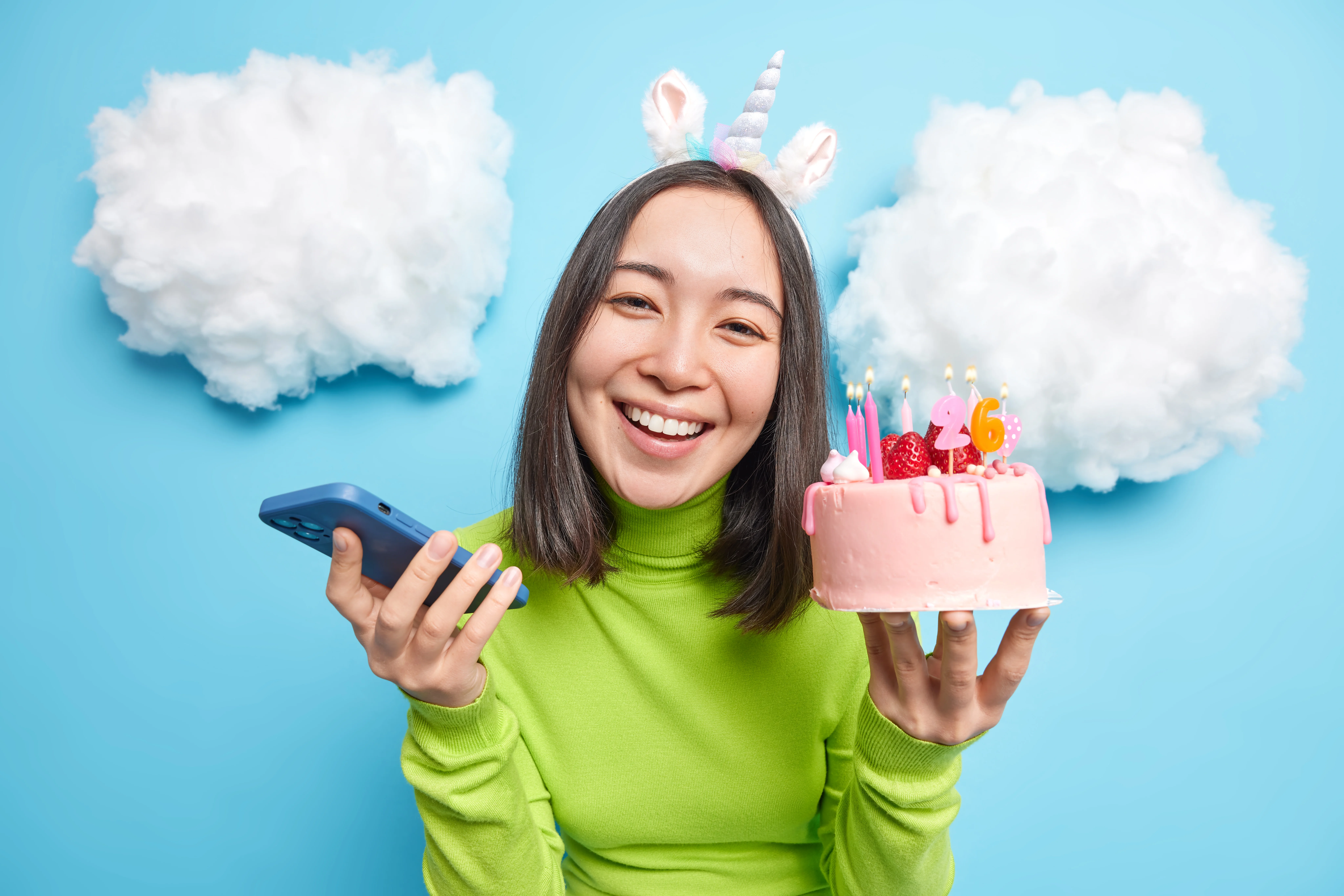 Smiling woman wearing a unicorn headband holds a pink birthday cake with candles showing the number 26 in one hand and a smartphone in the other, standing against a bright blue background with fluffy white cloud decorations.