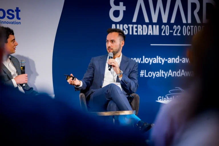 Ben Lipsey from Flying Blue speaking into a microphone while seated on stage at the Loyalty & Awards conference in Amsterdam. He is wearing a dark blue suit and looking toward another speaker seated to his right.