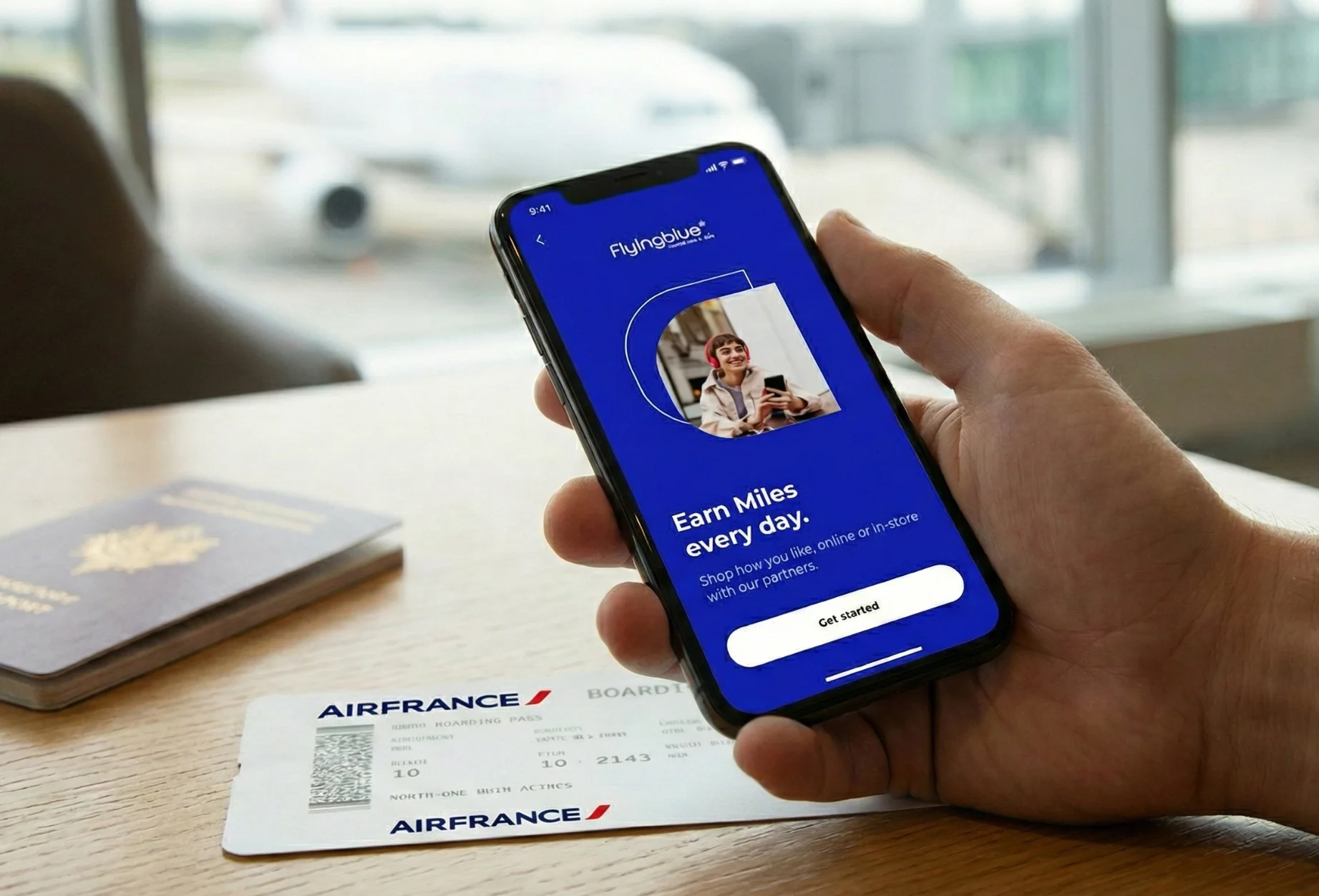 A person sitting at an airport lounge holding a smartphone displaying the Flying Blue app screen, which reads “Earn Miles every day.” A passport and a Flying Blue boarding pass lie on the table, with an airplane visible through the window in the background.